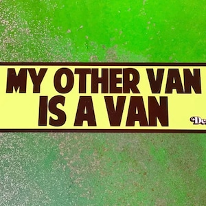 May include: A rectangular sticker with rounded corners, featuring the text "MY OTHER VAN IS A VAN" in dark brown letters on a yellow background. The sticker also includes the "Deathtrap" logo in the bottom right corner.