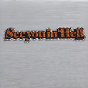 May include: A holographic sticker with the text "See you in Hell" in orange letters with a black outline. The sticker has a diamond pattern and a small "TheDaddy" logo in the bottom right corner.