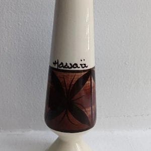 May include: A white ceramic vase with a brown and black geometric design. The vase has a flared base and a narrow neck. The word "Hawaii" is written on the vase in black.
