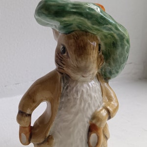 May include: A ceramic figurine of Peter Rabbit wearing a green hat with a brown jacket. The figurine is detailed with a white fur coat and brown eyes.