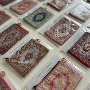Puede incluir: Una colección de estuches rectangulares con cremallera que presentan intrincados patrones de alfombras tejidas en varios colores, incluyendo rojo, azul, rosa y gris. Cada estuche está empaquetado individualmente en una funda de plástico transparente con una tarjeta que dice Thank you for celebrating Alisa's Ameen.