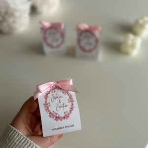 Può includere: Una piccola scatola bianca per bomboniere con un fiocco rosa, con un design a ghirlanda floreale con i nomi "Juliana & Jonathan" e la data "9 novembre 2025". La scatola è tenuta in mano.