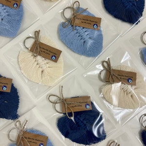 May include: A collection of macrame leaf keychains in cream, light blue, and dark blue, each individually packaged in clear plastic. Each keychain features a brown paper tag with the text Alp'in Mevlit Hatırası 14.06.2023 and a small evil eye bead.