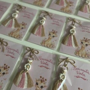 May include: Multiple macrame keychains featuring a small wooden flower bead and a multi-coloured tassel in beige, white, and pink. Each keychain is attached to a card with a giraffe illustration and the printed text Benvenuta Viola.