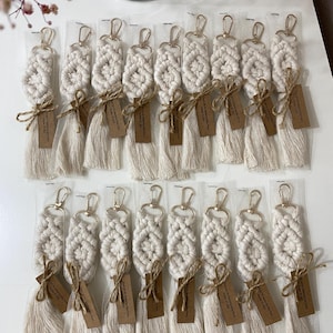Pode incluir: Uma coleção de dezoito porta-chaves de macramé em tom esbranquiçado, cada um com um padrão de nós, um fecho mosquetão dourado e uma borla comprida. Cada porta-chaves está embalado individualmente numa manga transparente com uma etiqueta de papel castanho.