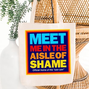 以下が含まれることがあります： 「Meet me in the aisle of shame. Official home of the 'caw caw'」と書かれたカラフルなグラフィックが施された白いキャンバス地のトートバッグ