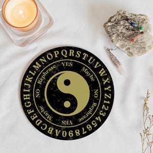 Puede incluir: Una tabla de Ouija negra y dorada con un símbolo yin yang en el centro. La tabla tiene letras, números y las palabras "Sí", "No", "Tal vez" y "Reformular" alrededor del borde.