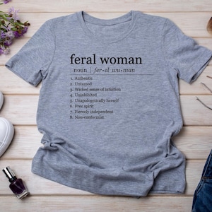 May include: Gray t-shirt with the text "feral woman" defined as "fer-el wu-man" and a list of eight characteristics: Authentic, Untamed, Wicked sense of intuition, Uninhibited, Unapologetically herself, Free spirit, Fiercely independent, Non-conformist.