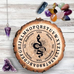 Op de afbeelding: Houten Ouija-bord met een zwarte slang en een bloemenpatroon. Het bord heeft letters, cijfers en de woorden "Ja", "Nee", "Misschien" en "Herformuleren".