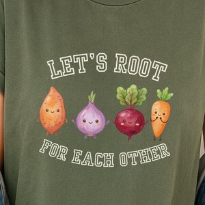 Op de afbeelding: Een militair groen t-shirt met een afbeelding van vier getekende wortelgroenten, waaronder een zoete aardappel, ui, biet en wortel. De tekst "LET'S ROOT FOR EACH OTHER" is in witte letters boven en onder de groenten gedrukt.