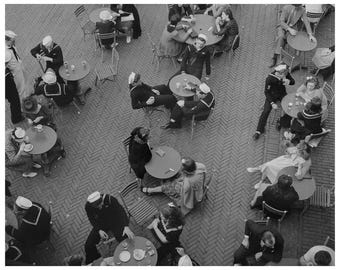 May include: A high-angle black and white photograph of a crowded outdoor cafe with a herringbone brick floor. Numerous individuals are seated at small round tables, many wearing military-style uniforms and white sailor caps.