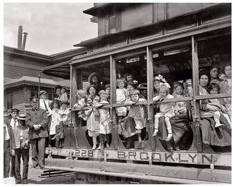 May include: A black and white photograph of a trolley car labeled 228 BROOKLYN filled with children and adults. A uniformed conductor stands on the ground beside the vehicle, while passengers look out from the open-sided wooden carriage.