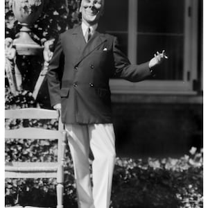 May include: A man wearing a white mask with a wide smile, a dark suit jacket, white pants, and black shoes. He is standing in front of a window and a potted plant.