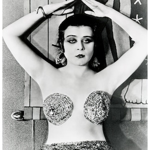 May include: Black and white vintage photograph of a woman with dark hair and makeup, arms raised above her head. She wears decorative, circular pasties and a sequined skirt. The image has a classic, old Hollywood aesthetic.