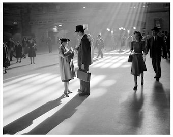 May include: Black and white photograph of a train station scene. Several people are walking in the station. A man in a suit and hat stands with a woman in a coat and hat. Other people are walking in the background.