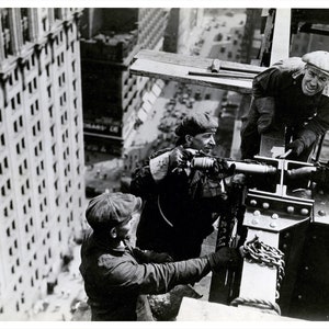 May include: Black and white photograph of construction workers on a high-rise building. Three men are working on a steel beam, with one using a drill. The image captures the dangerous work environment and the height of the building.