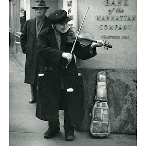 May include: A man plays a violin on the street in front of a bank. The bank is called "Bank of the Manhattan Company" and has the year "1799" on it. A second man in a coat stands behind the violinist.