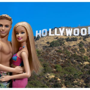 May include: A couple of dolls, a man and a woman, are embracing in front of the Hollywood sign. The woman doll has long blonde hair and is wearing a pink and blue swimsuit. The man doll is shirtless and wearing patterned shorts. The Hollywood sign is white and set against a blue sky.