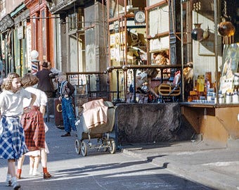 May include: A vintage street scene featuring a sidewalk with people walking past storefronts. A baby carriage sits near a shop entrance with a Coca-Cola sign. Store windows display various goods, including a sign that reads Prescriptions 598.