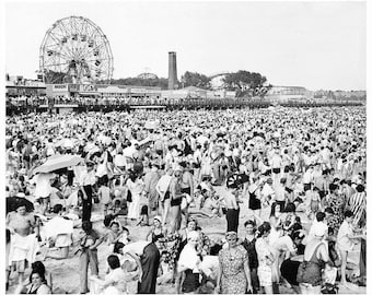 May include: A black and white photograph of a crowded beach scene at Coney Island. A large Ferris wheel stands in the background above a boardwalk with various buildings. Numerous people are gathered on the sand in swimwear and summer clothing.
