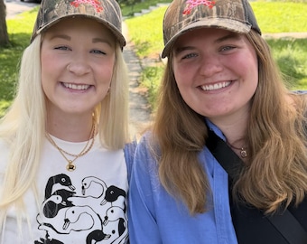 May include: Two baseball caps featuring a camouflage pattern in shades of brown, green, and black. Each cap displays a small, embroidered graphic of a pink horse with red spots centered on the front panel above the curved brim.