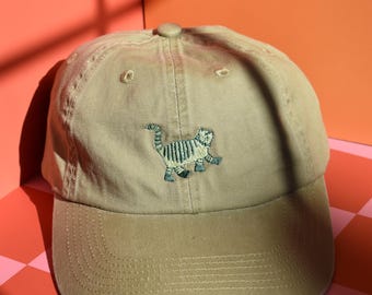 May include: A tan baseball cap featuring an embroidered graphic of a small, striped cat in shades of green and grey. The cap has a structured crown, a curved brim, and a matching fabric button at the top center, with visible stitching details.
