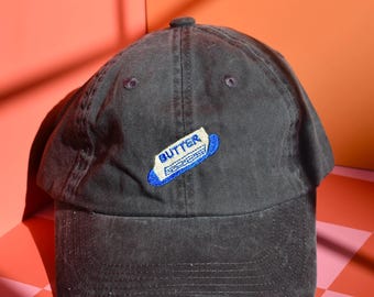 May include: A charcoal grey baseball cap featuring an embroidered patch on the front. The patch depicts a stick of butter with the word BUTTER stitched in blue letters on a white background, outlined in a vibrant blue border.