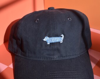 May include: A black baseball cap featuring an embroidered light blue dachshund dog with white striped detailing on its body. The cap has a curved brim, a button on the top, and stitched eyelets for ventilation, providing a structured look.
