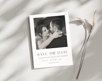 May include: A black and white save-the-date card featuring a romantic photo of a couple embracing. The card includes the text "SAVE THE DATE" and details: Alex and Olivia are getting married August 27th, 2027, in Cleveland, OH. Dried flowers are in the background.