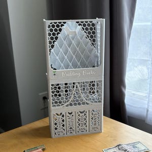 May include: A gray, 3D-printed money organizer with the text "Building Bucks." It has compartments labeled "Charity," "Save & Invest," "Needs," and "Wants," with percentage allocations. Rolls of coins and US dollar bills are on the wooden surface.