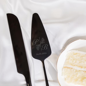 May include: Black cake knife and server set with engraved initials "P B" and the date "04.05.2026". The server has a serrated edge. A slice of cake is visible on a white plate.