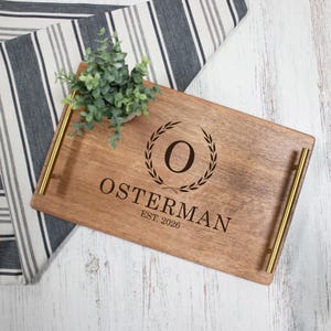 May include: A rectangular wooden serving tray with gold-toned metal handles. The surface features a laser-engraved design with a central letter O surrounded by a laurel wreath, the text OSTERMAN, and the text EST. 2026 below the name.