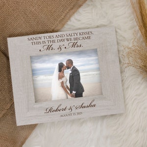 May include: A light gray wooden picture frame with a wedding photo. The frame has the text "SANDY TOES AND SALTY KISSES, THIS IS THE DAY WE BECAME Mr. & Mrs." and the names "Robert & Sasha" with the date "AUGUST 12, 2023".