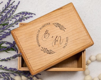 May include: A wooden box with a lid slightly ajar, revealing an empty interior. The lid is engraved with the words "WHISPERS OF FOREVER", initials "B + M", and the date "10.22.2025" within a circular wreath design. The box is surrounded by lavender and white beads.