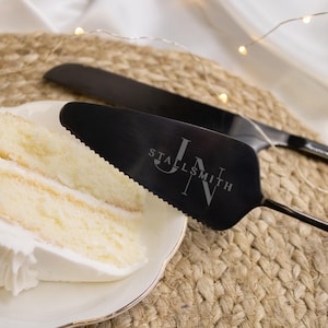 May include: A black cake server and knife set with the initials "JN" and the last name "STALLSMITH" engraved on the server. The server has a serrated edge. A slice of cake with white frosting sits on a white plate.