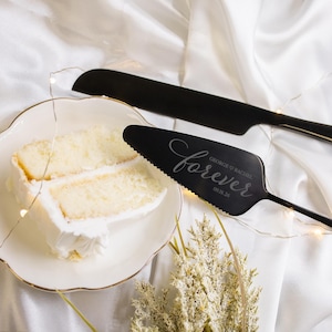 May include: A black cake knife and server set with the word "forever" engraved on the server, along with names and a date. A white cake with frosting sits on a white plate with a gold rim. The background is white fabric.