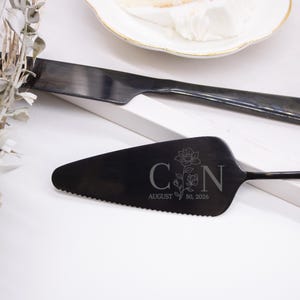 May include: Black cake knife and server set. The server is engraved with the initials "C & N", a floral design, and the date "August 30, 2026". The knife and server are placed near a white plate with a slice of cake.