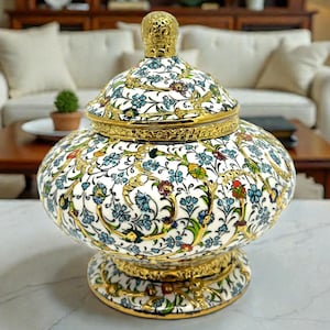 Puede incluir: Un tarro decorativo de cerámica con tapa. La base blanca está adornada con un estampado floral en azul, verde y rojo. Tiene detalles dorados y una tapa a juego con un remate. Un elemento decorativo para el hogar.