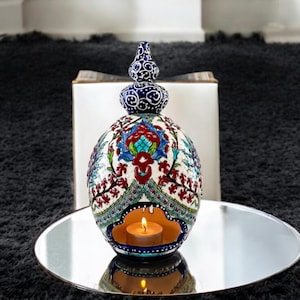 May include: A white ceramic candle holder with blue and red floral designs. The holder has a round shape with a small opening at the top for a tea light candle. The candle holder is sitting on a silver tray.