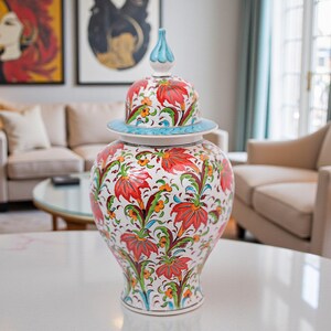 Op de afbeelding: Een decoratieve keramische gemberpot met deksel, met een levendig bloemmotief in rood, oranje en groen op een witte achtergrond. De pot heeft een lichtblauwe rand en een bijpassende knop op het deksel. Een decoratief woondecoratie-item.