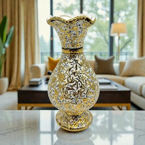 Könnte beinhalten: Eine dekorative Vase mit einem weißen und goldenen Blumendesign. Die Vase hat einen gerüschten oberen Rand und einen bauchigen Körper. Die goldenen Akzente und komplizierten Muster schaffen eine elegante Ästhetik. Die Vase steht auf einer Marmoroberfläche.