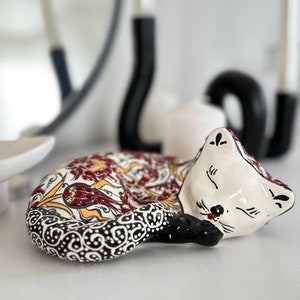 May include: A white ceramic cat figurine with red, yellow, and black painted details. The cat is curled up in a sleeping position.