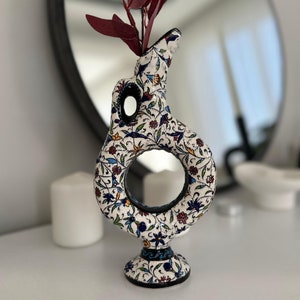 Może przedstawiać: Biały ceramiczny wazon z kwiatowym wzorem i okrągłym otworem. Wazon ma okrągłą podstawę i jest ozdobiony niebieskimi, zielonymi, czerwonymi i żółtymi kolorami.