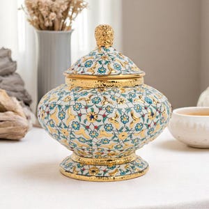 Könnte beinhalten: Dekorative Keramikdose mit Deckel, verziert mit einem Blumenmuster in Blau, Gold und Rot auf weißem Grund. Die Dose hat goldene Ränder und einen goldenen Knauf auf dem Deckel. Die Dose steht auf einer weißen Oberfläche.