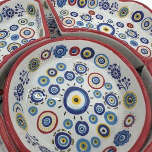 Peut inclure: Un ensemble de plats de service en c&eacute;ramique avec un fond blanc et une bordure rouge. Les plats sont d&eacute;cor&eacute;s de motifs d'&oelig;il mal&eacute;fique bleus, jaunes et rouges.