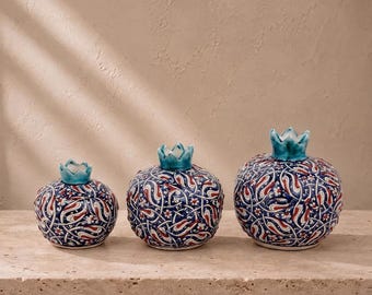 May include: Three ceramic pomegranate-shaped decorative objects in varying sizes. Each piece features a dark blue base with intricate white and red floral patterns and a glossy turquoise crown-like top, arranged in a row on a flat surface.