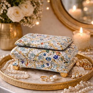 May include: A rectangular white ceramic jewelry box featuring a detailed floral pattern in blue, green, and orange with gold-toned vine accents. The box has a hinged lid and sits on four ornate gold-toned feet, resting on a round gold tray.