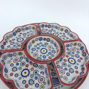 Peut inclure: Un plat de service en c&eacute;ramique blanche avec un bord rouge et un motif d'&oelig;il mal&eacute;fique bleu et jaune. Le plat est divis&eacute; en six sections, avec un petit bol au centre.
