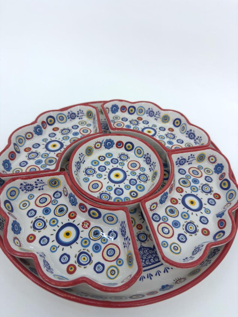 Peut inclure: Un plat de service en c&eacute;ramique blanche avec un bord rouge et un motif d'&oelig;il mal&eacute;fique bleu et jaune. Le plat est divis&eacute; en six sections, avec un petit bol au centre.