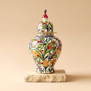 Può includere: Un vaso in ceramica con coperchio rimovibile su base bianca, decorato con motivi floreali dipinti a mano nei toni del rosso, giallo, verde e blu. Il vaso presenta un corpo arrotondato, un collo affusolato e un pomello appuntito sul coperchio.